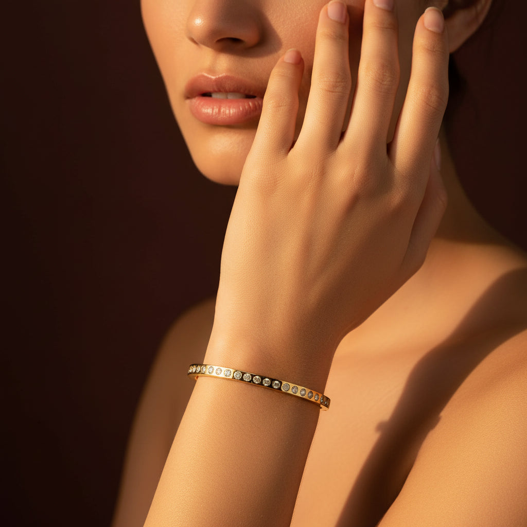 A close-up flat lay photograph focusing tightly on a model's hand and wrist wearing the standard size gold crystal eternity kada under warm golden lighting against a rich #5b3b3a brown background. Face half visible.