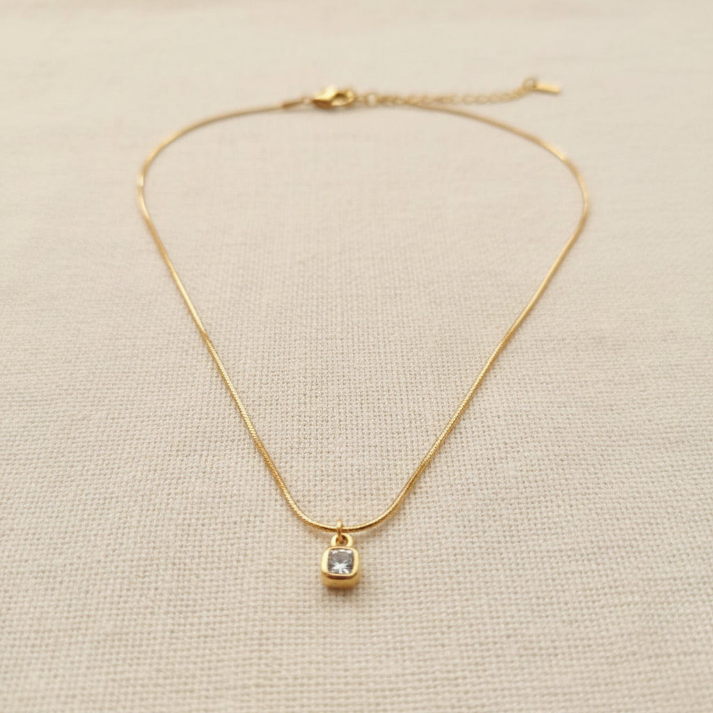 A realistic photograph of the fluid 18K gold snake chain cushion pendant necklace resting naturally on a textured cream linen cloth under natural light.
