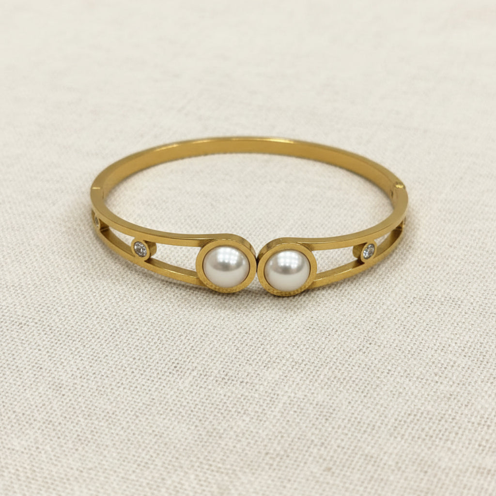 A realistic, natural daylight flat lay photograph of the premium gold plated open cutout double pearl statement bracelet resting naturally on a textured cream linen cloth.