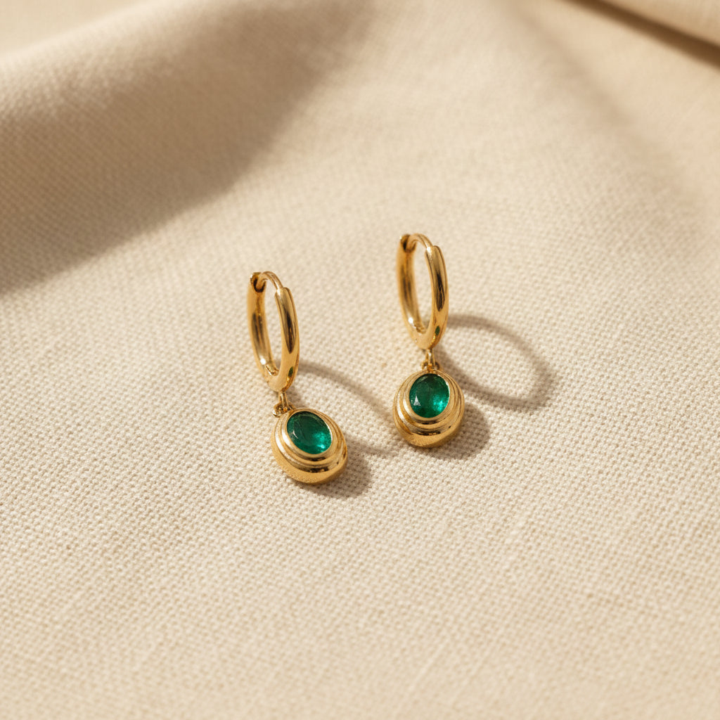 Realistic flat lay shot of the gold oval emerald drop huggie earrings resting on a textured cream linen cloth under natural lighting.
