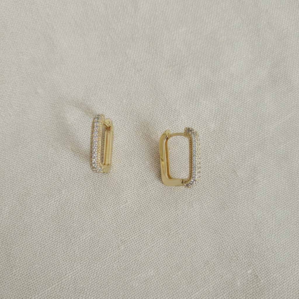 Realistic flat lay shot of the gold rectangular crystal huggie earrings resting on a textured cream linen cloth under natural lighting.
