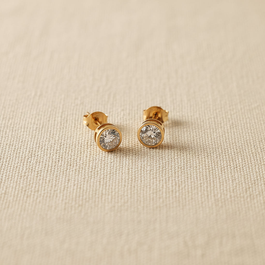 Realistic flat lay shot of the gold solitaire crystal stud earrings resting on a textured cream linen cloth under natural lighting.