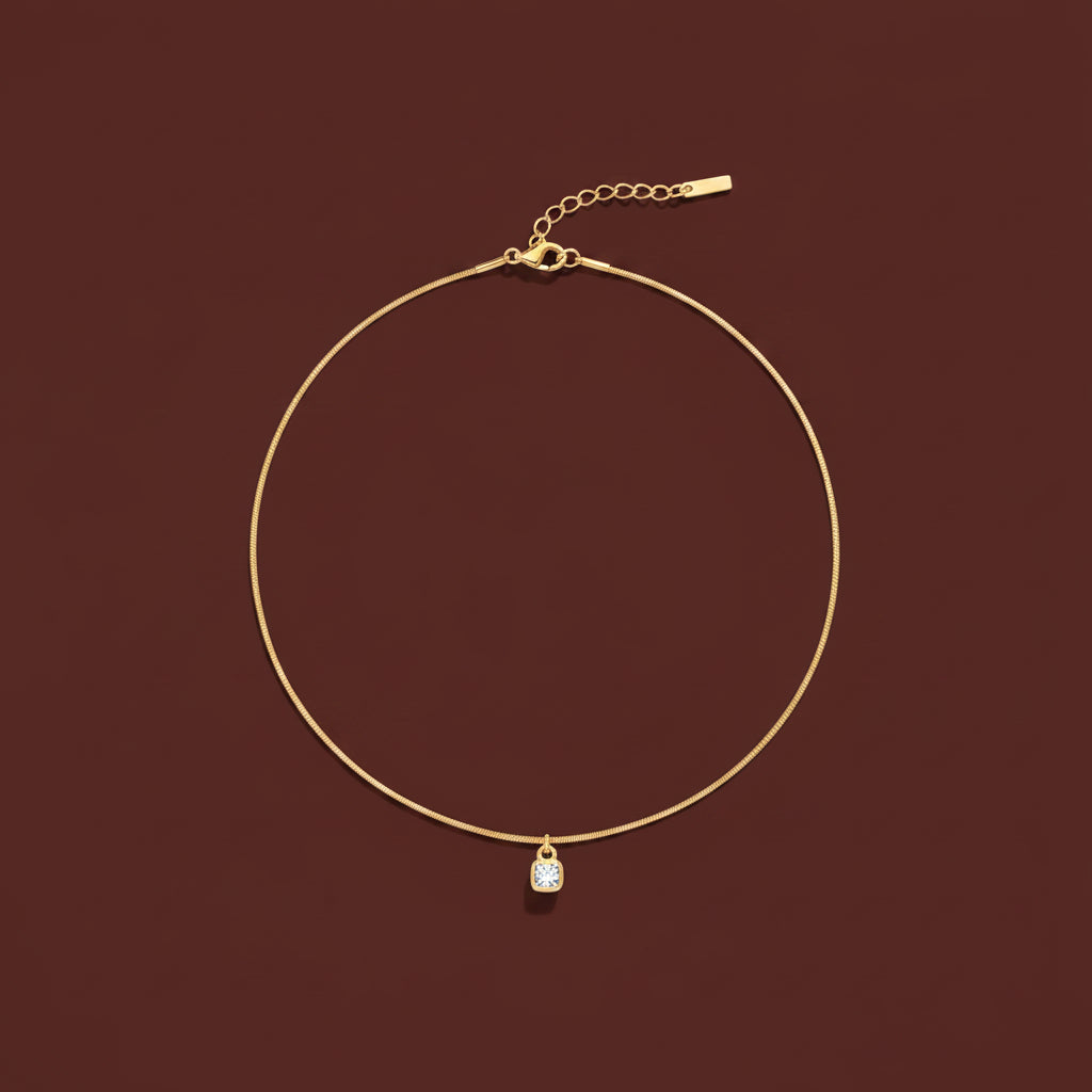 A professional flat-lay catalogue image showing the 18K gold cushion-cut crystal bezel necklace arranged in a precise circle with soft shadows on a rich cocoa brown background.