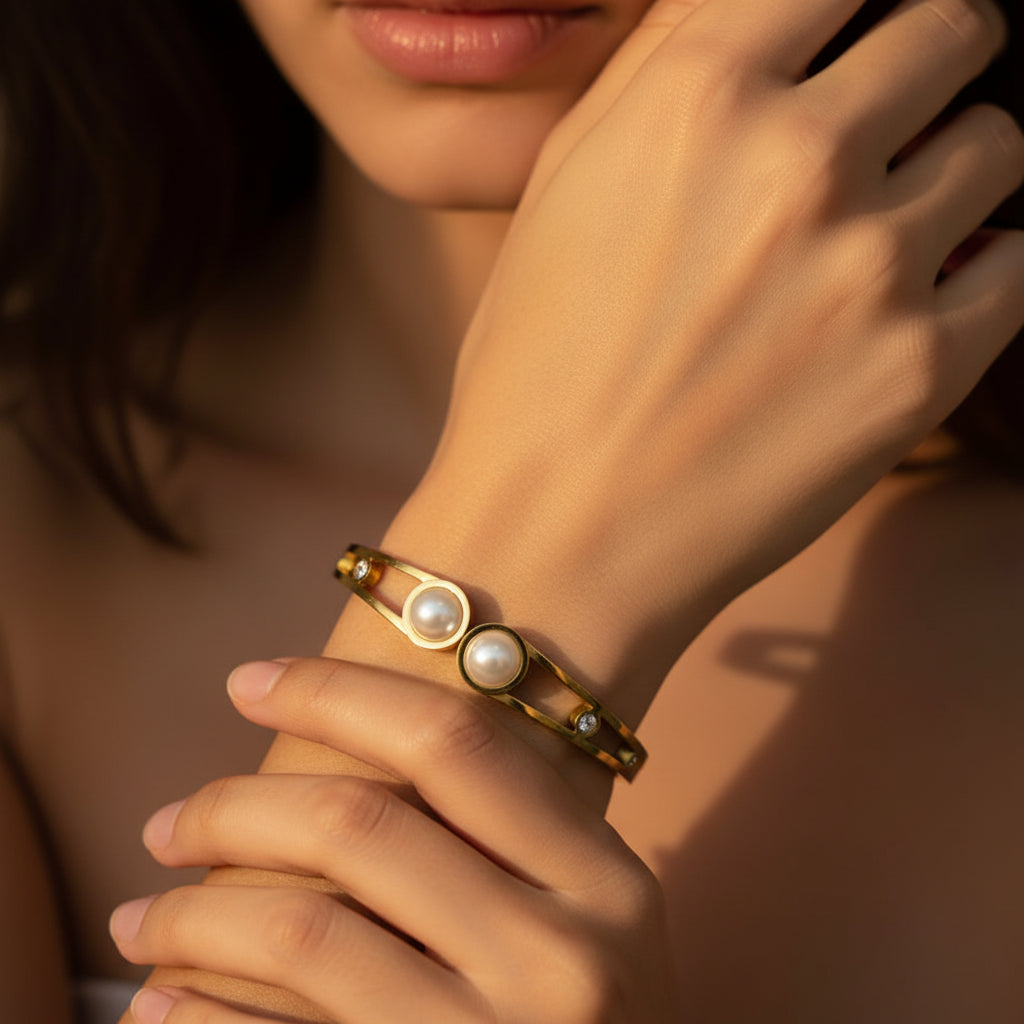 A close-up flat lay photograph focusing closely on a model's hand and wrist wearing the standard size gold open split-band double pearl kada under warm golden lighting against a rich #5b3b3a brown background. Face half visible.