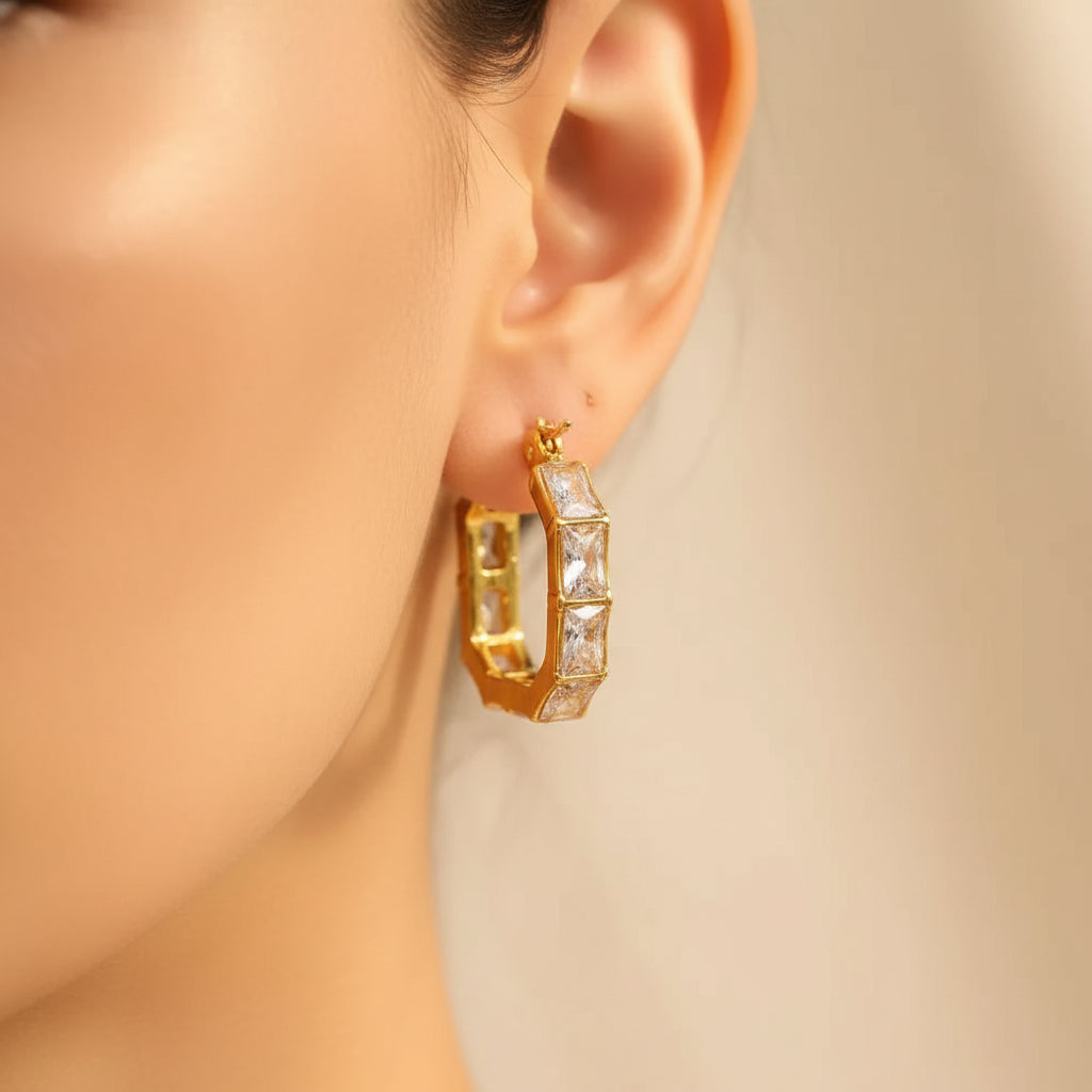 Zoomed-in flat shot focusing closely on a model's ear wearing the gold hexagon hoop earrings lined with square-cut stones under warm golden lighting against a rich #5b3b3a brown background.