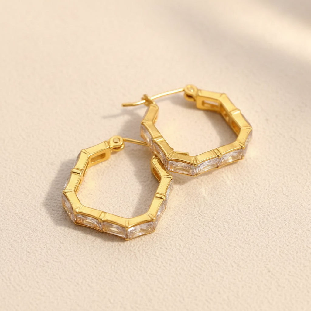 A professional standing catalogue shot of the 18K gold plated geometric hexagon hoop earrings with square crystals with simple shadows on a light #f5f3eb cream background.