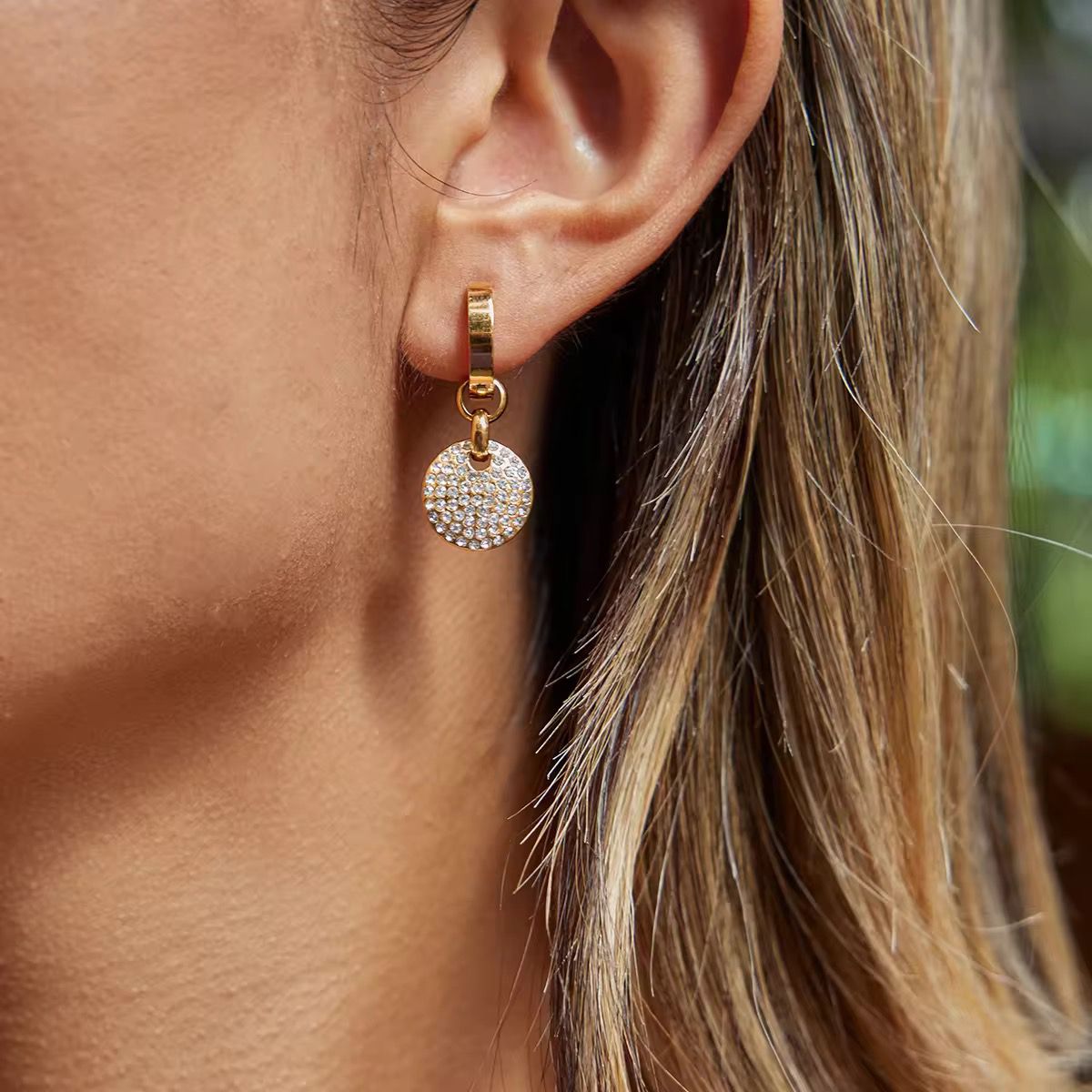 Zoomed-in lifestyle view of a model wearing the gold circular micro-pavecrystal disc drop earrings in warm golden lighting against a rich brown background.