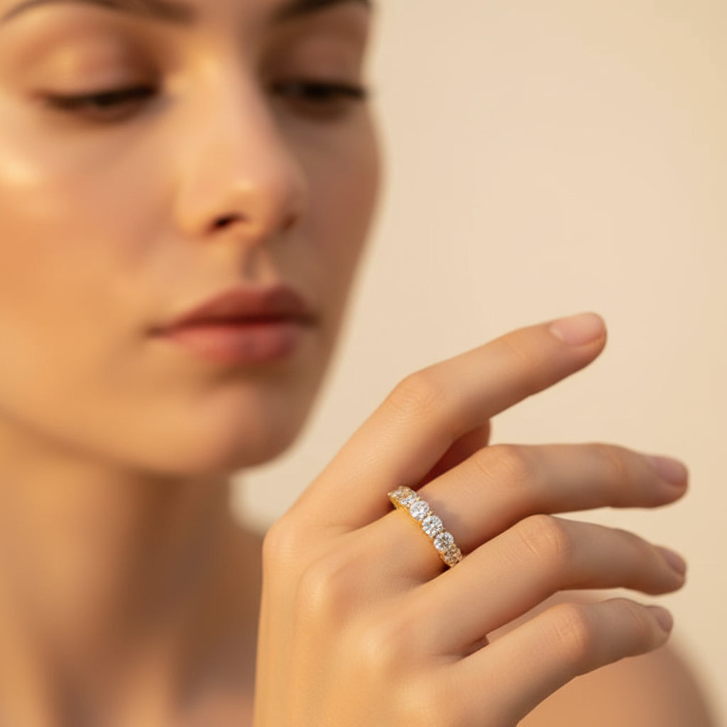 A close-up flat photograph of a model with her face half-visible, showcasing the gold seven-stone crystal half eternity ring on her hand under warm golden lighting against a light #f5f3eb cream background.