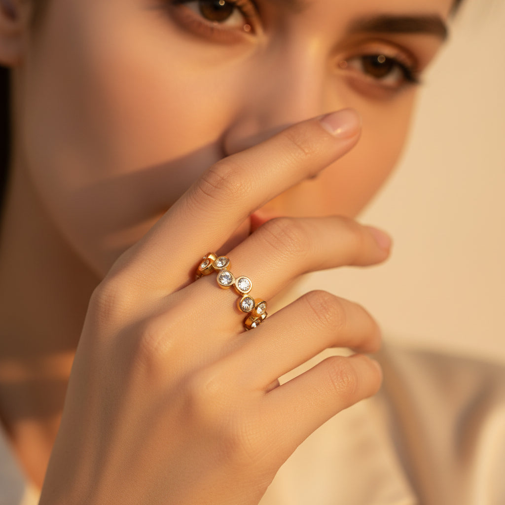 Close-up flat photograph of a model with her face half-visible, showcasing the gold interlocking crystal eternity ring under warm golden lighting against a cream background.