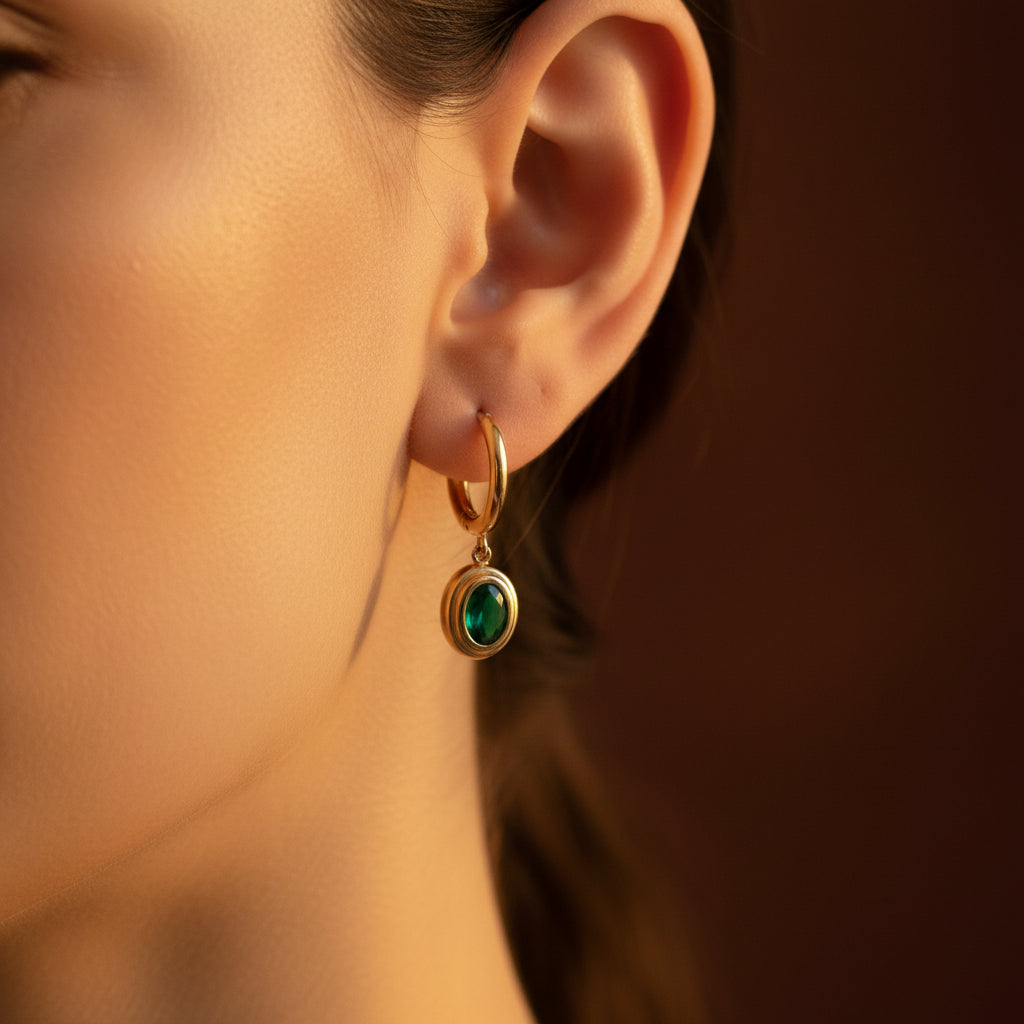 Zoomed-in lifestyle view of a model wearing the gold green emerald drop huggie earrings in warm golden lighting on a rich brown background.
