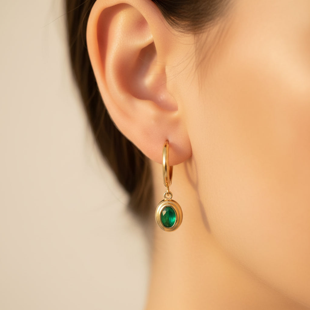 Close-up side profile of a model wearing the gold oval emerald drop huggie earrings under warm golden lighting on a minimal cream background.