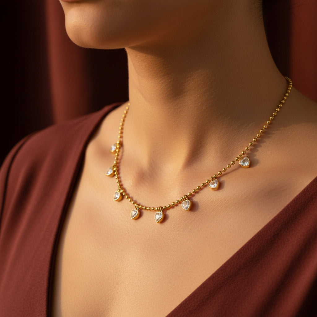 A detailed, realistic photograph focusing closely on a woman's neck, wearing the 18K gold plated heart crystal droplet station ball chain necklace under warm golden lighting against a solid deep cocoa brown background.