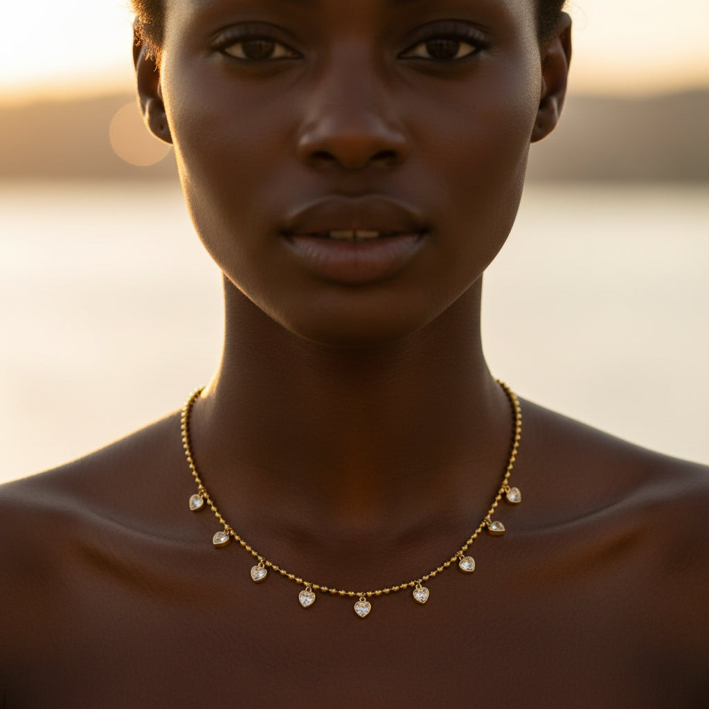 A stunning high-end e-commerce editorial photograph of a beautiful Black model wearing the gold cascading heart crystal ball chain necklace under warm golden lighting.