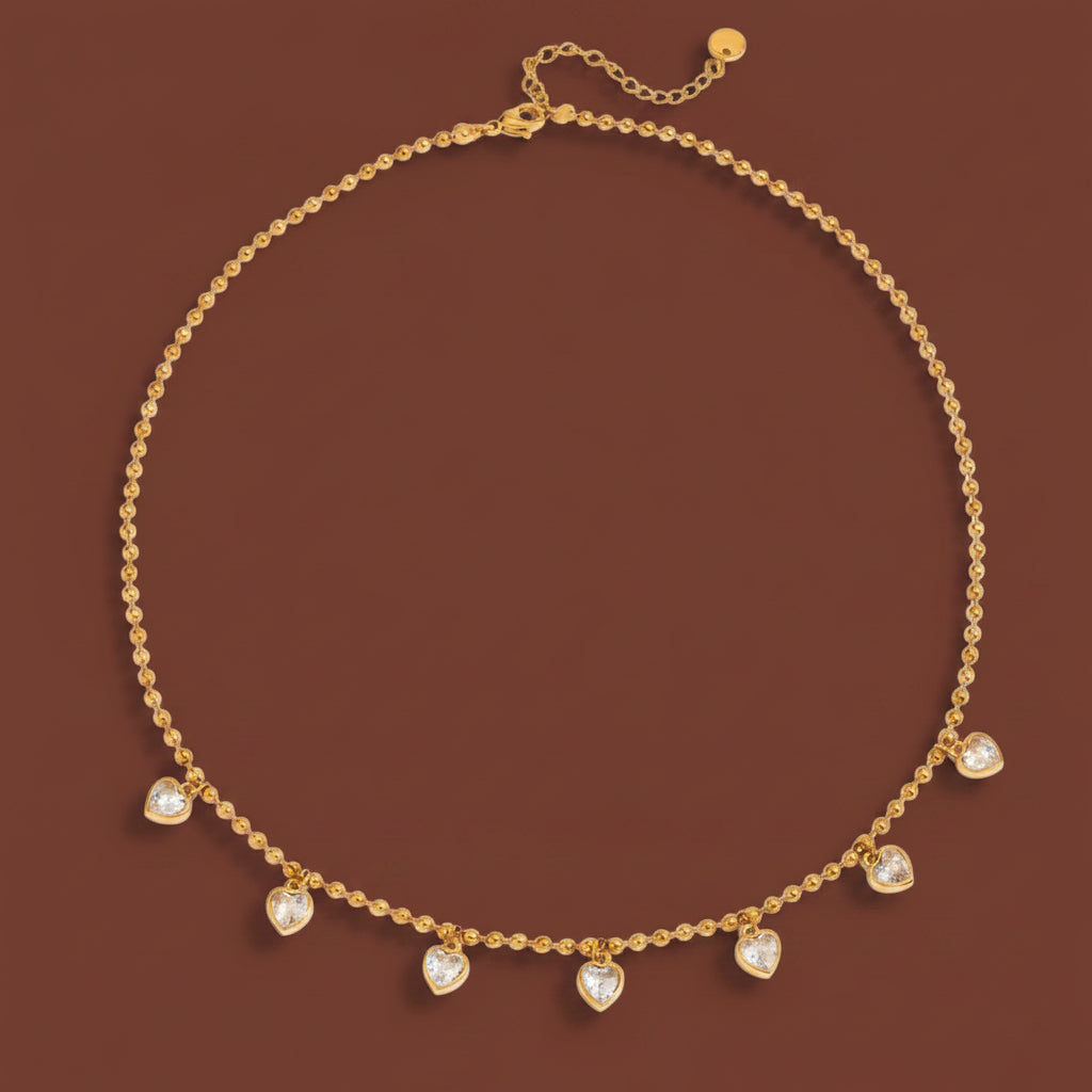 A professional catalogue flat-lay image showing the 18K gold micro-ball chain necklace with hanging heart crystal charms arranged in a precise circle with clean shadows on a plain cocoa brown background.