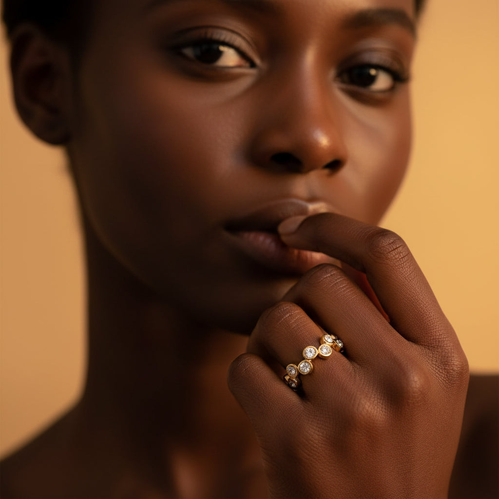 A stunning high-end e-commerce editorial photograph of a beautiful Black model under warm golden lighting, with her face visible and the focus zoomed in tightly on the gold crystal zig-zag ring.