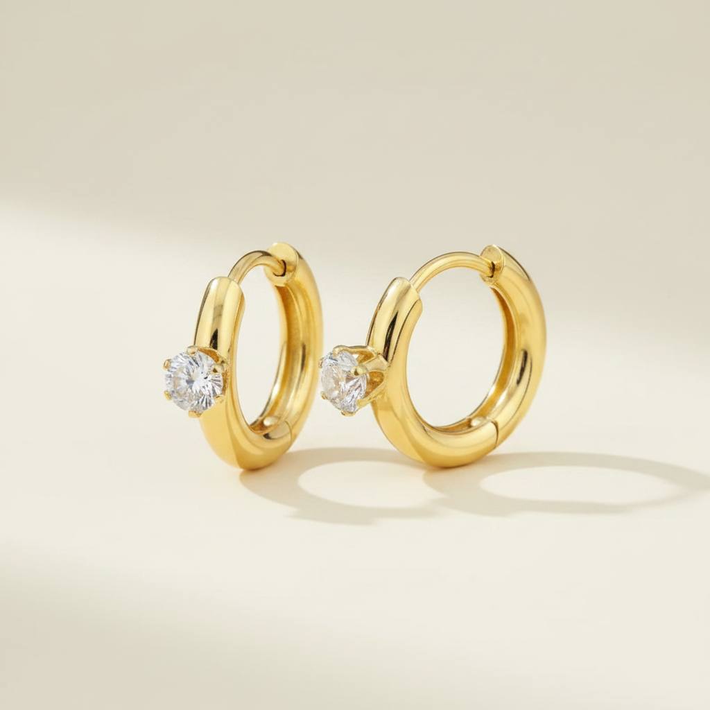 Gold solitaire huggie earrings on a premium cream catalogue background.