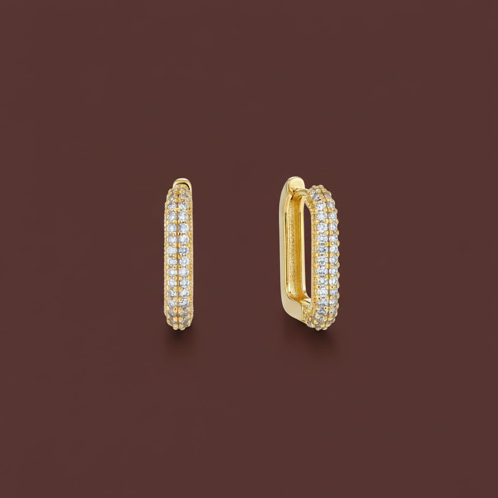 Professional catalogue flat lay of Verge rectangular crystal huggie earrings in 18K gold plating with soft shadows on a rich brown background.