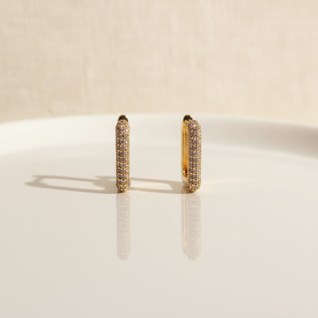 Professional standing catalogue shot of the 18K gold plated geometric crystal hoop earrings with a simple shadow on a light cream background.