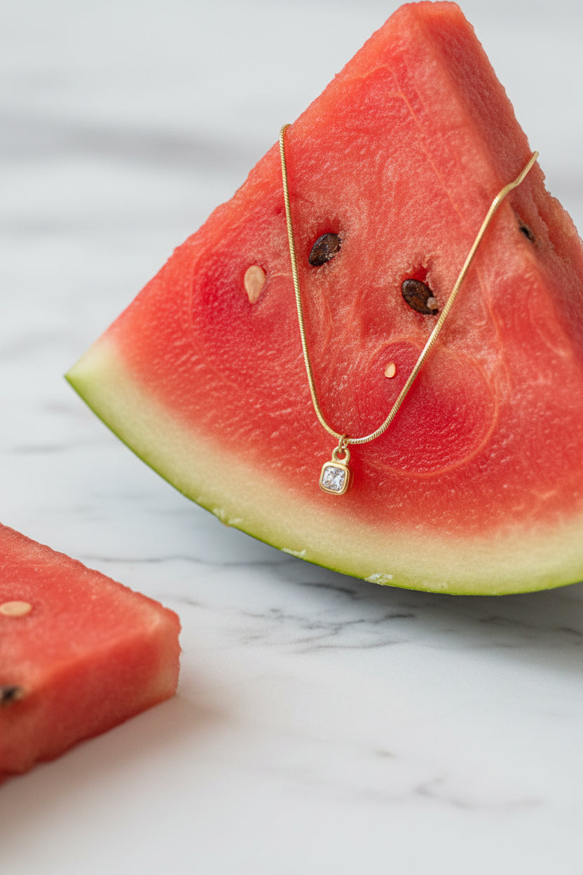 A high-end editorial lifestyle photograph capturing the cushion-cut solitaire bezel necklace composed appealingly on a refreshing food item, with sharp focus on the central square gemstone pendant.