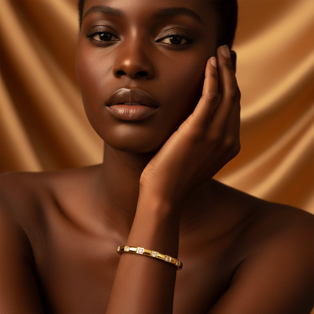 A stunning high-end e-commerce editorial photograph of a beautiful Black beauty model under warm golden lighting, with her face fully visible and the focus zoomed in tightly on the small size square-framed crystal gold oval kada bracelet on her slender wrist.