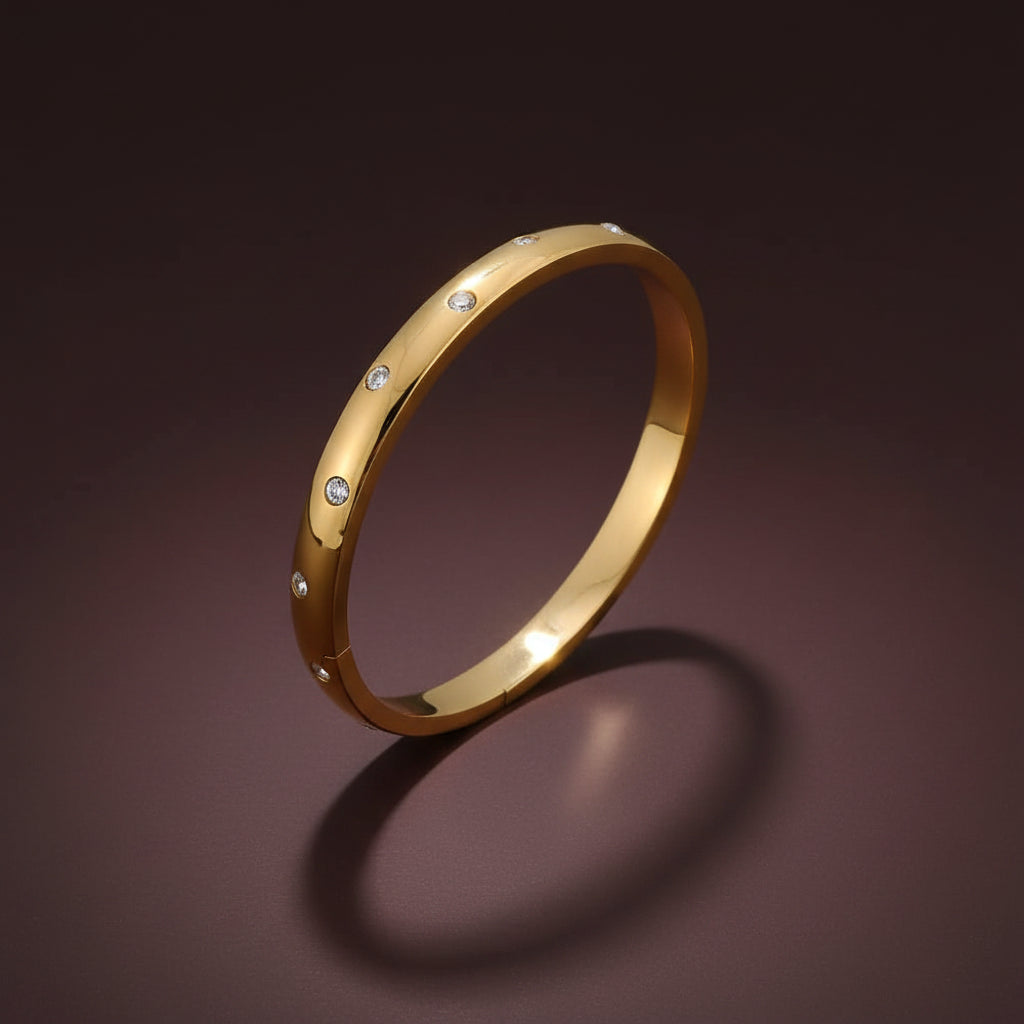 A professional high-angle catalogue shot of the dainty oval gold plated stainless steel kada bangle with embedded crystals, specifically sized for thin wrists, isolated on a deep cocoa brown background with clean shadows.