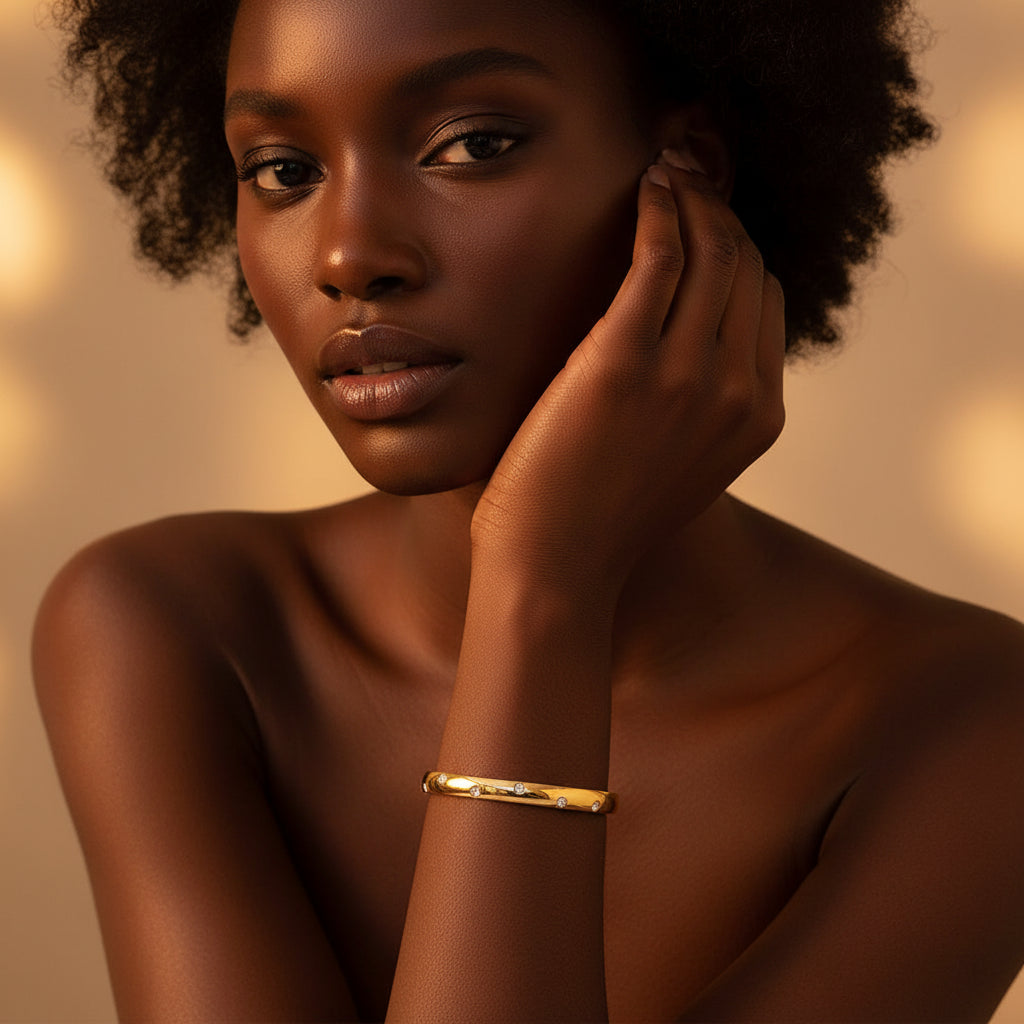 A stunning high-end e-commerce editorial photograph of a beautiful Black beauty model under warm golden lighting, with her face fully visible and the focus zoomed in tightly on the small size gold crystal oval kada bracelet on her slender wrist.