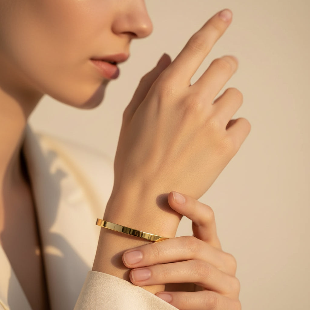 A standing profile photograph of a model (face half visible), showcasing the smooth small size gold oval plain bracelet on her thin wrist under warm golden lighting against a cream background, as seen in IMG_5657.webp.