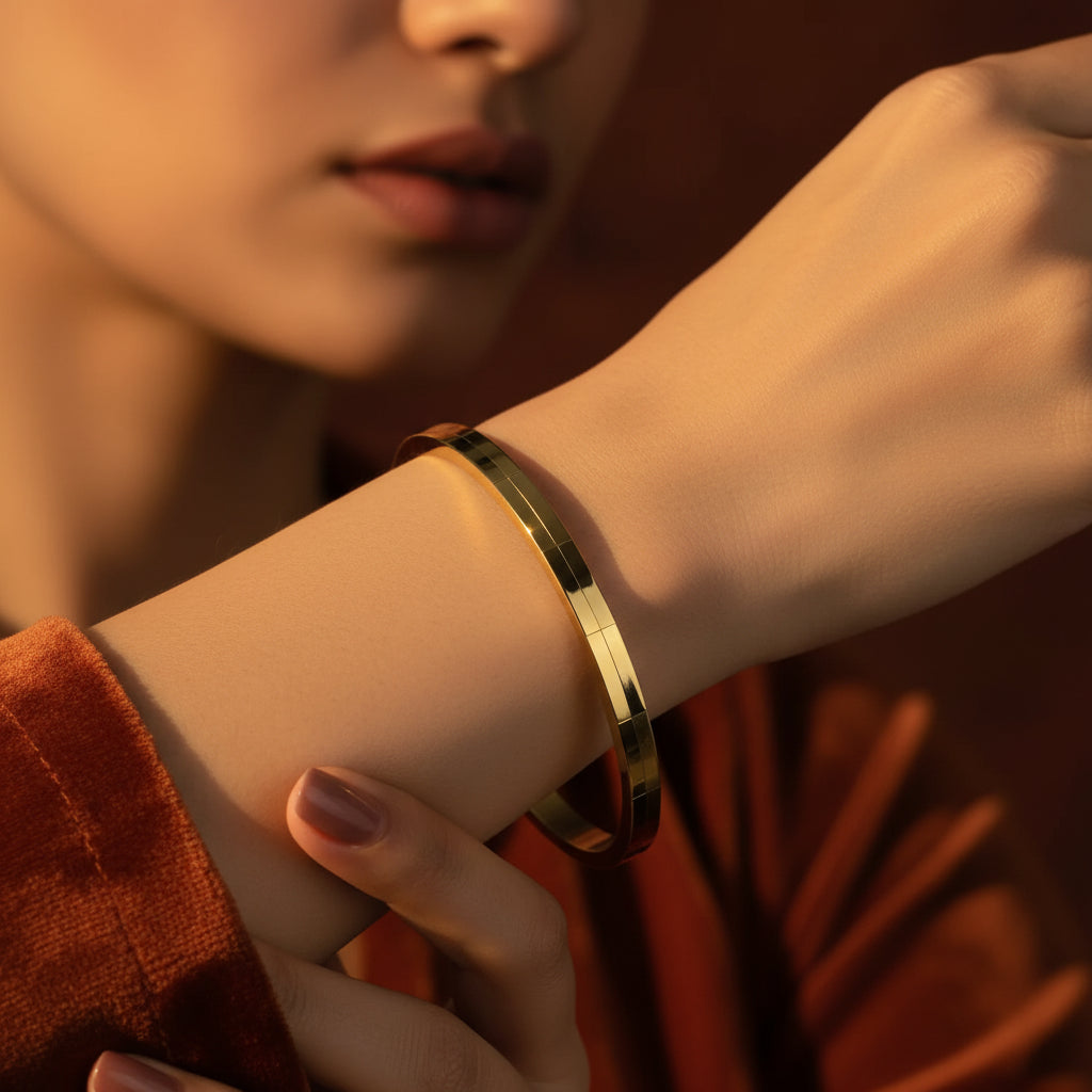 A close-up flat lay photograph focusing closely on a model's distinctly slender, thin hand and wrist wearing the small oval brick grid textured gold kada under warm golden lighting against a deep brown background.