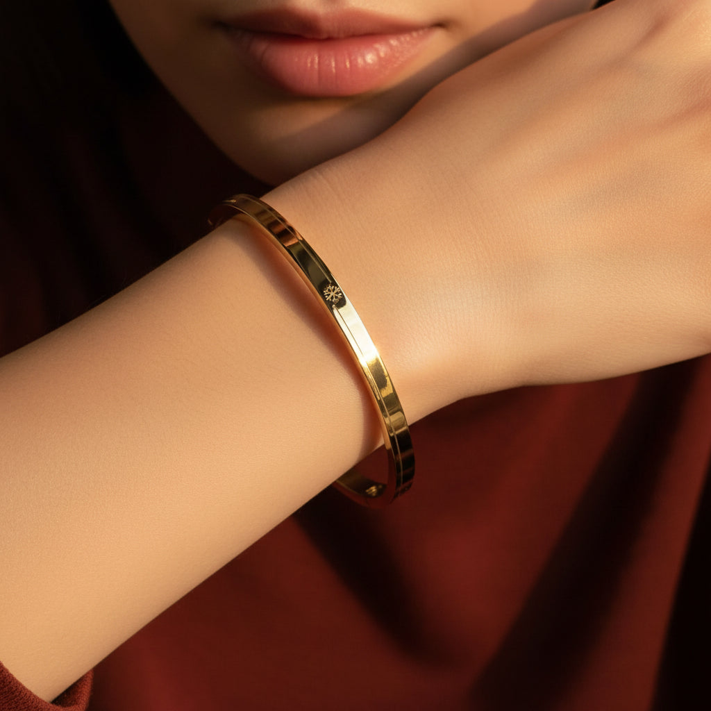 A close-up flat lay photograph focusing closely on a model's distinctly slender, thin hand and wrist wearing the small oval snowflake engraved gold kada under warm golden lighting against a deep brown background.