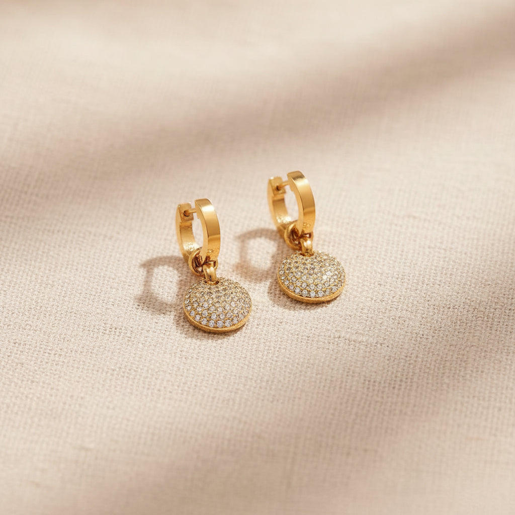 Realistic flat lay shot of the premium gold round pavé crystal charm drop earrings resting beautifully on a textured cream linen cloth.