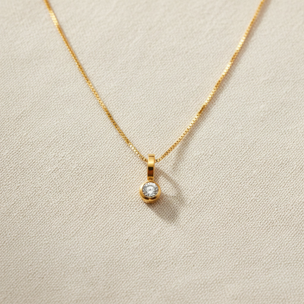 A realistic photograph of the ultra-fine 18K gold round solitaire pendant necklace resting naturally on a textured cream linen cloth under natural light.