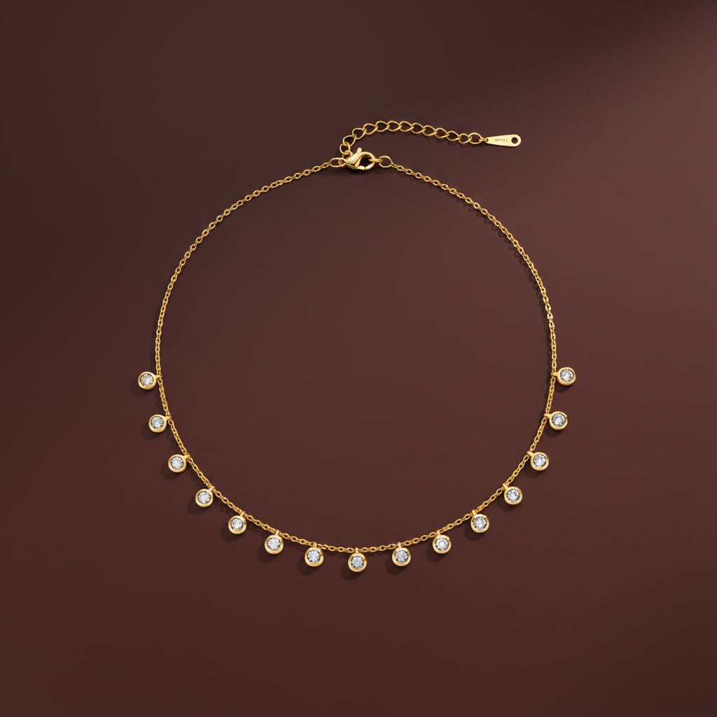 A professional catalogue flat-lay image showing the 18K gold micro-ball chain necklace with hanging crystal stations arranged in a precise circle with clean shadows on a plain cocoa brown background.