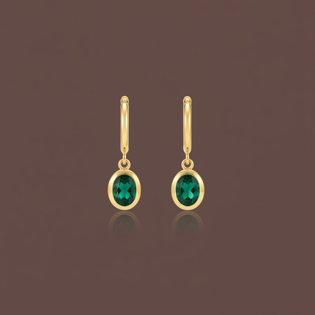 Professional catalogue flat lay of Verdant oval emerald drop huggie earrings in 18K gold plating with soft shadows on a rich brown background.