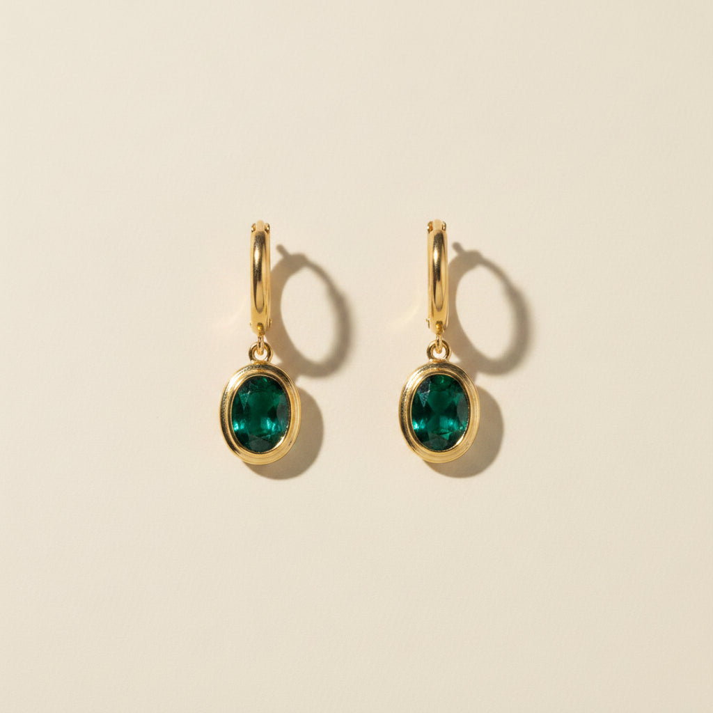 Professional standing catalogue shot of the 18K gold plated oval green gemstone hoop earrings with a simple shadow on a light cream background.