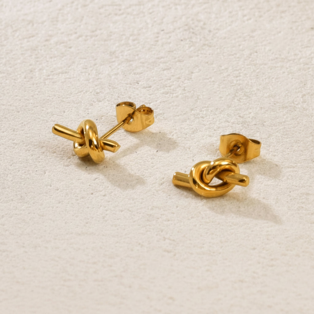 Gold knot stud earrings on a premium #f5f3eb catalogue background.
