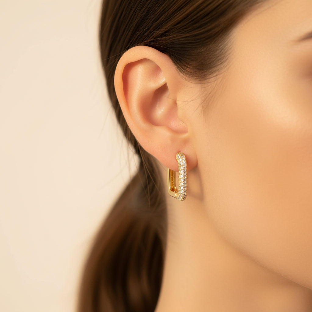 Close-up side profile of a model wearing the gold rectangular crystal huggie earrings under warm golden lighting on a minimal cream background.