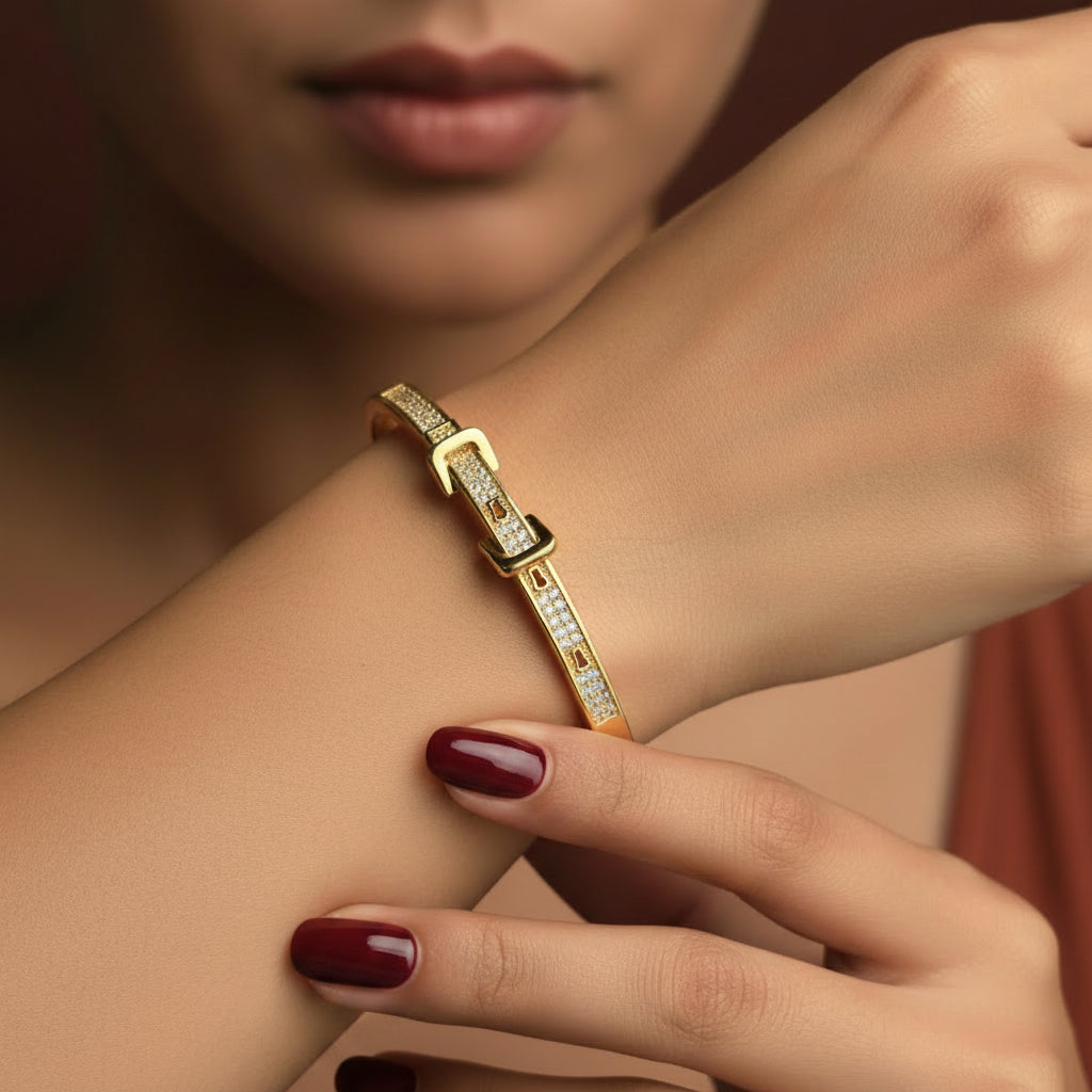 A close-up flat lay photograph focusing closely on a model's hand and wrist wearing the standard size gold belt buckle crystal kada under warm golden lighting against a rich #5b3b3a brown background.
