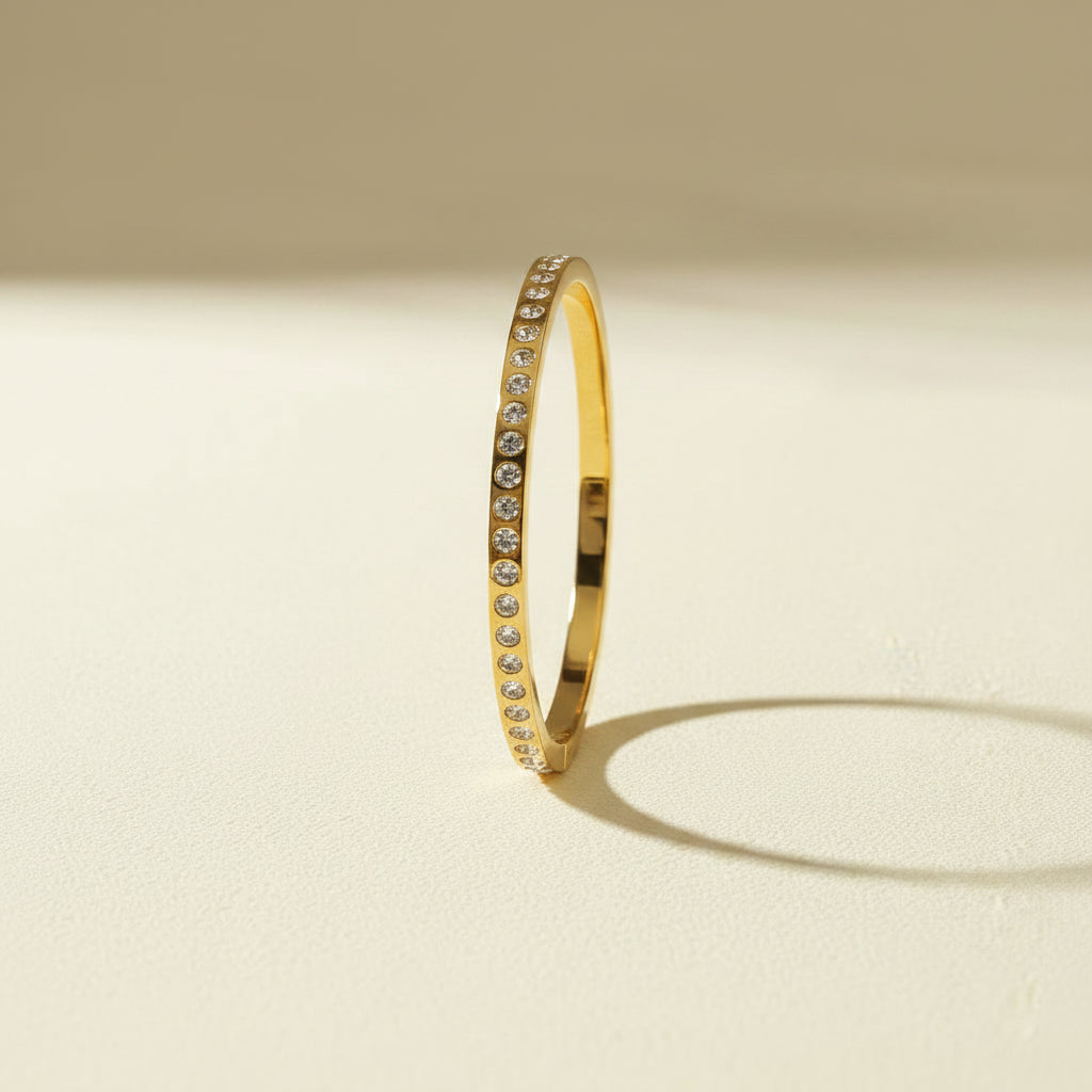 A professional standing catalogue view of the regular size 18K gold plated continuous crystal eternity bangle with simple shadows on a light #f5f3eb cream background.