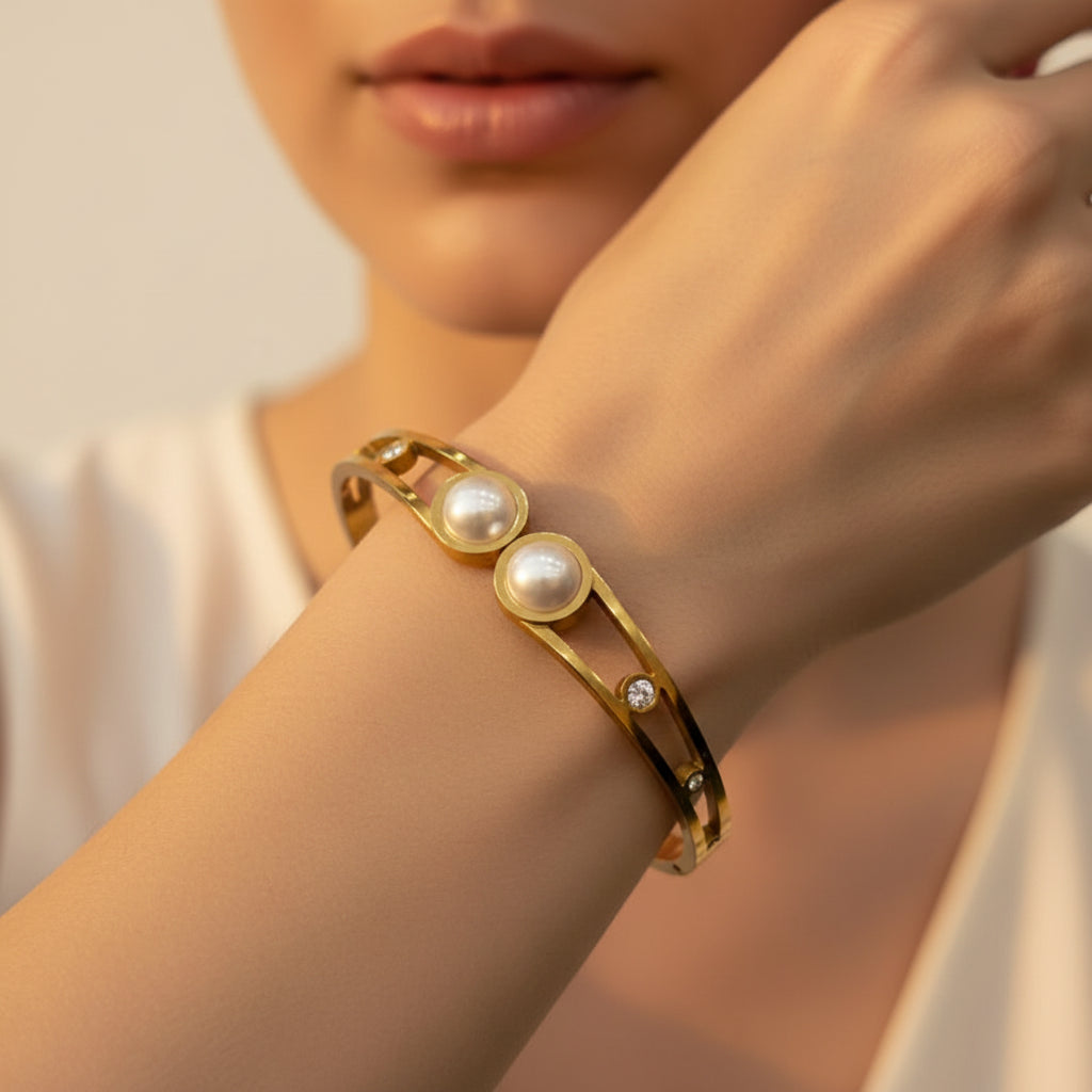 A standing profile photograph of a model (face half visible), highlighting the split open-frame double pearl gold bracelet on her wrist under warm golden lighting against a light #f5f3eb cream background.