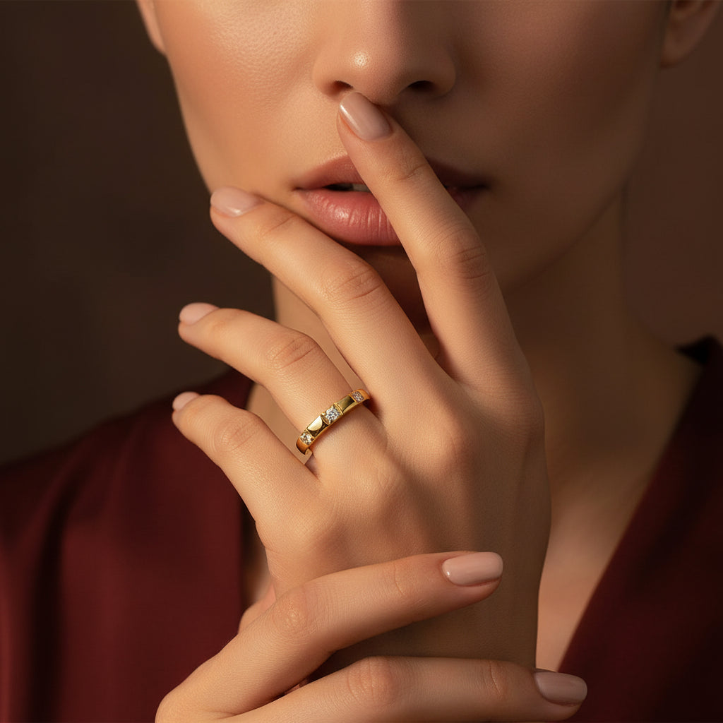 Zoomed-in flat layout of a model with her face half-visible, highlighting the gold flat-edge triple stone stacker ring on her finger under warm golden lighting against a rich #5b3b3a brown background.