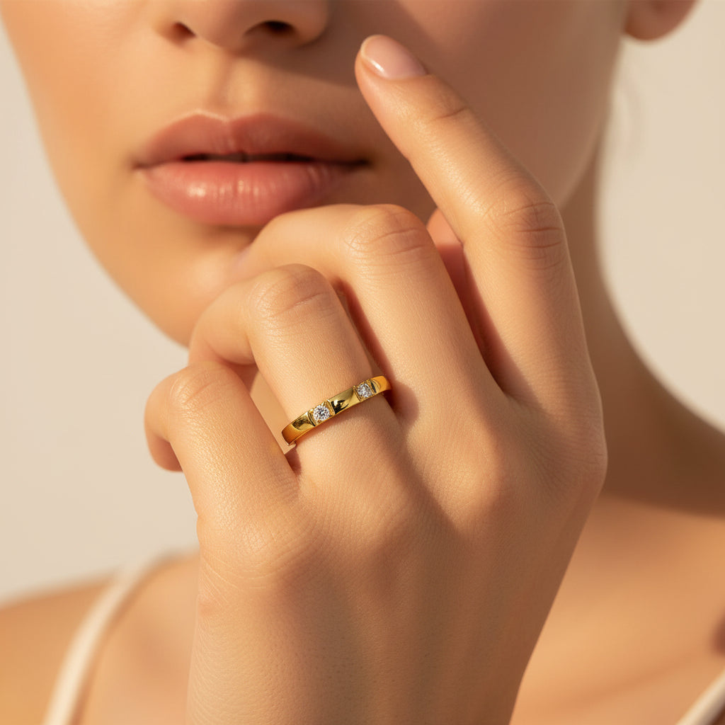 A close-up flat photograph of a model with her face half-visible, showcasing the gold flat-edge triple stone stacking ring on her hand under warm golden lighting against a light #f5f3eb cream background.