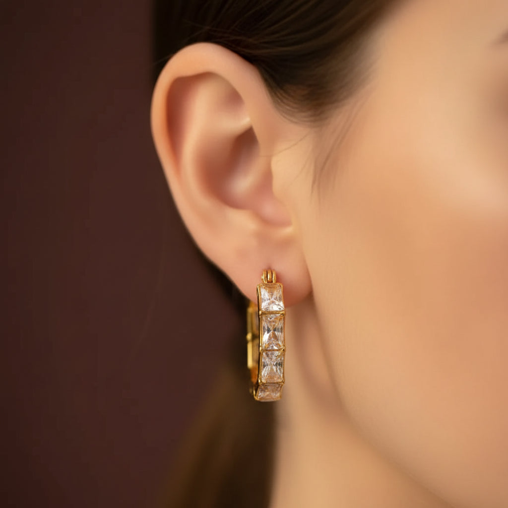 A close-up flat photograph focusing closely on a model's ear wearing the gold six-sided crystal hoop earrings under warm golden lighting against a light #f5f3eb cream background.