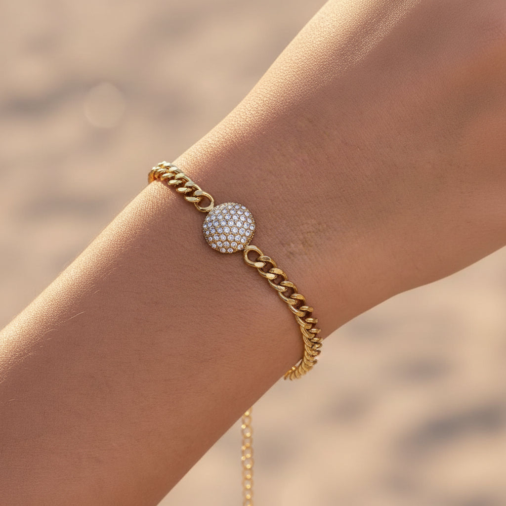 A detailed, realistic photograph focusing closely on a woman's wrist wearing the gold micro-pavé crystal disc curb chain bracelet under warm golden lighting.