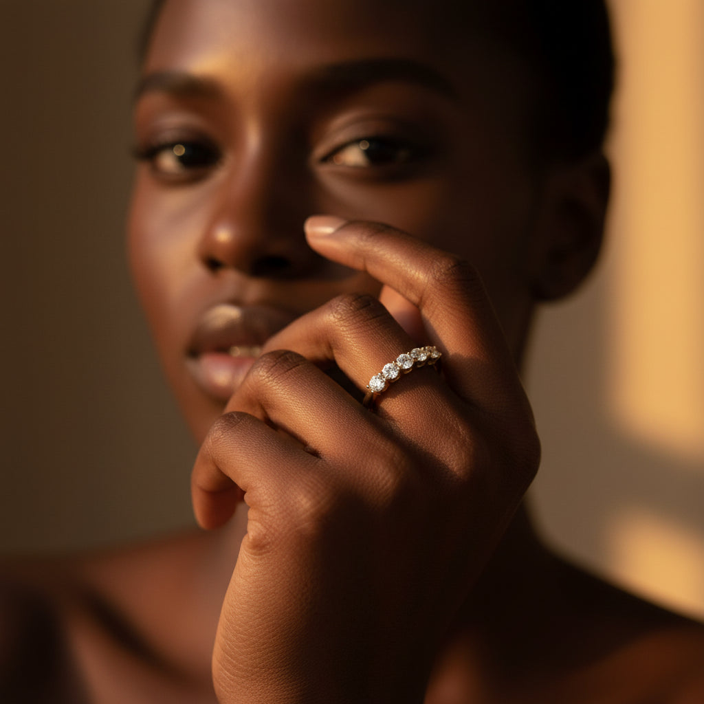 A stunning high-end e-commerce editorial photograph of a beautiful Black beauty model under warm golden lighting, with her face visible and the focus zoomed in tightly on the gold seven stone shared prong crystal ring without changing its design.