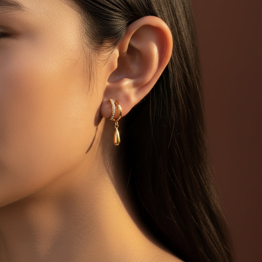 Zoomed-in lifestyle shot of a model with her face half-visible, showcasing the gold split double-hoop crystal teardrop earrings against a rich brown background.