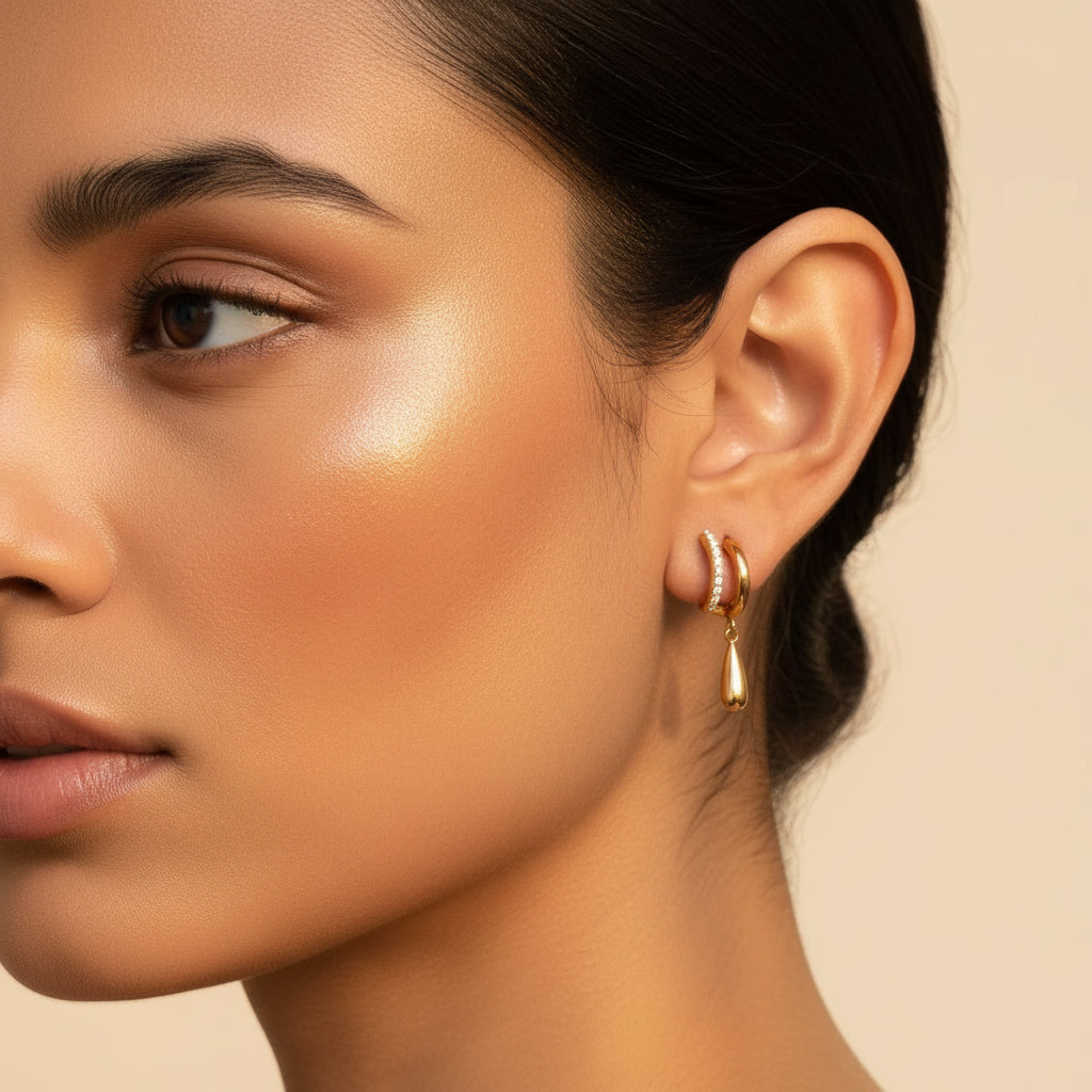 A standing profile photograph of a model with her face half-visible, highlighting the radiant gold split-hoop crystal teardrop earrings under warm golden lighting against a cream background.