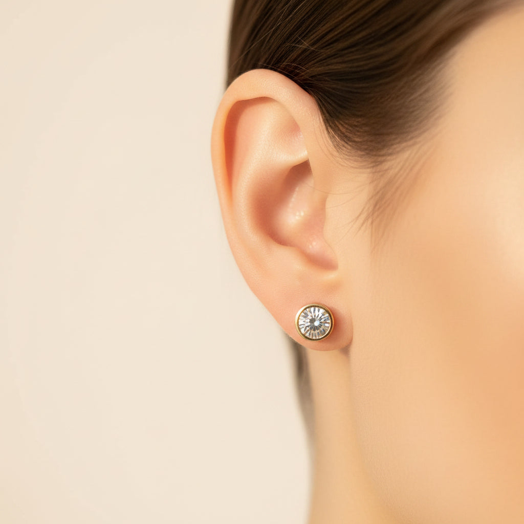 Close-up side profile of a model wearing the gold crystal bezel stud earrings under warm golden lighting on a minimal cream background.