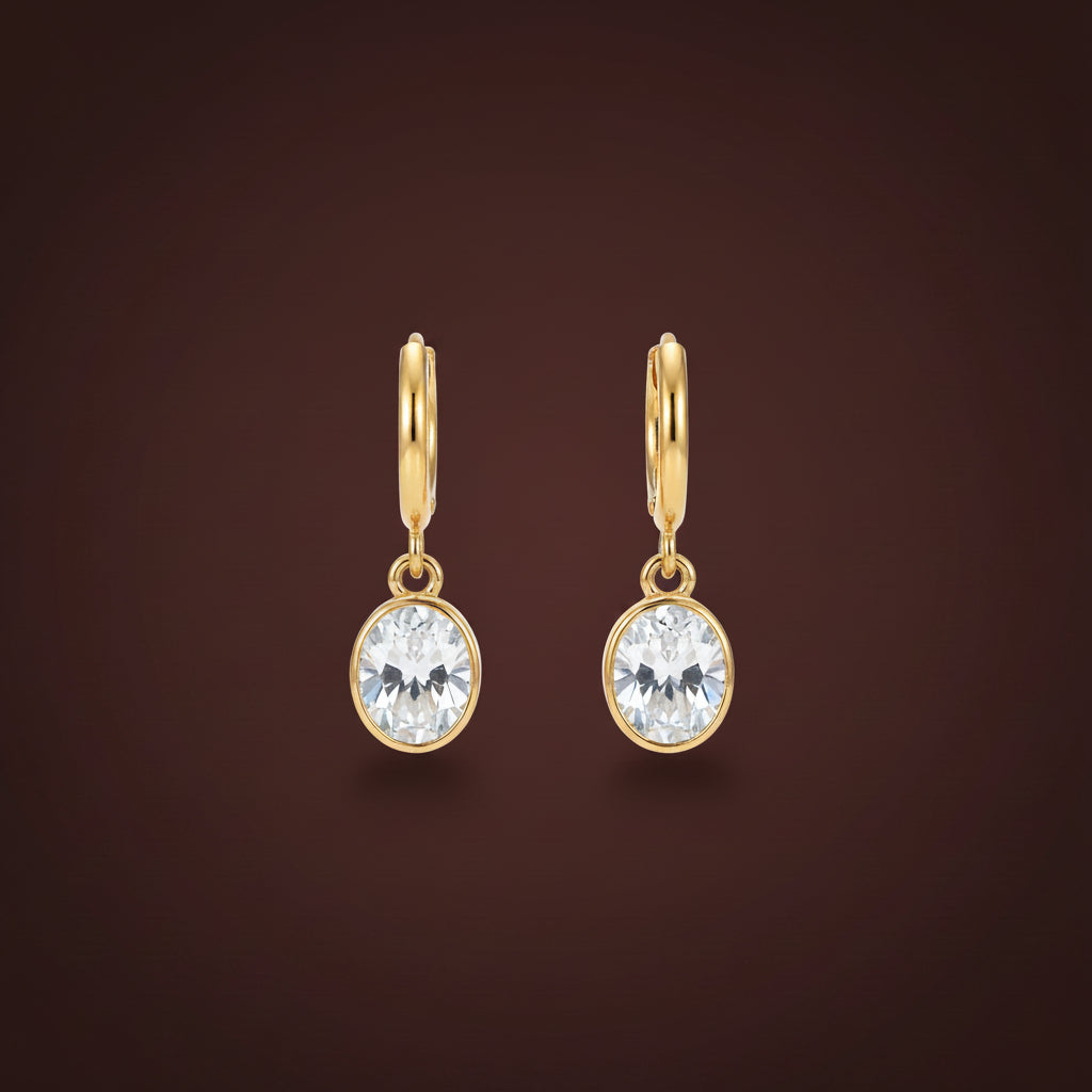 Front view of Aura oval crystal drop huggie earrings in 18K gold plating on a premium display stand.