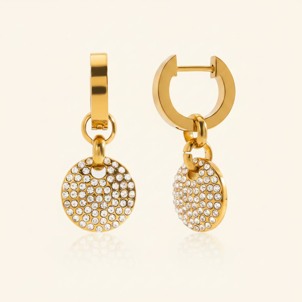 Professional standing catalogue shot of the 18K gold plated sparkling circle pendant huggie earrings with a simple shadow on a light cream background.