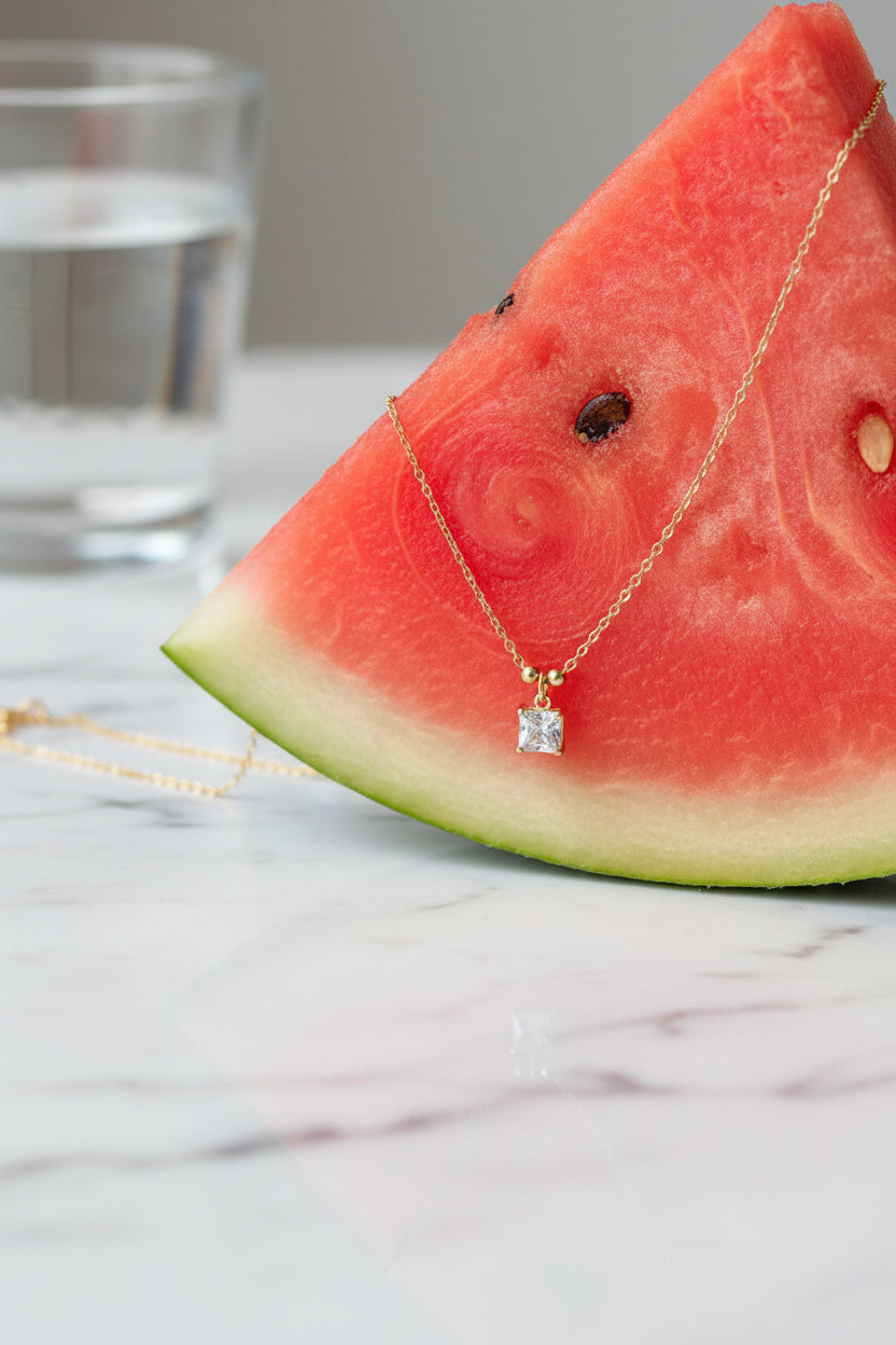 A high-end editorial lifestyle photograph capturing the princess-cut solitaire necklace composed appealingly on a refreshing food item, with sharp focus on the square crystal pendant.